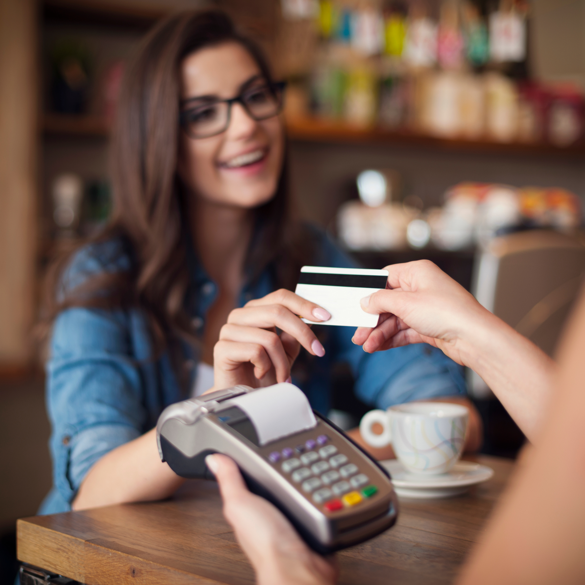 mulher sorrindo entregando cartão de crédito/débito sorrindo; mão esquerda de outra pessoa segurando máquina de cartão e mão direita pegando o cartão da mulher